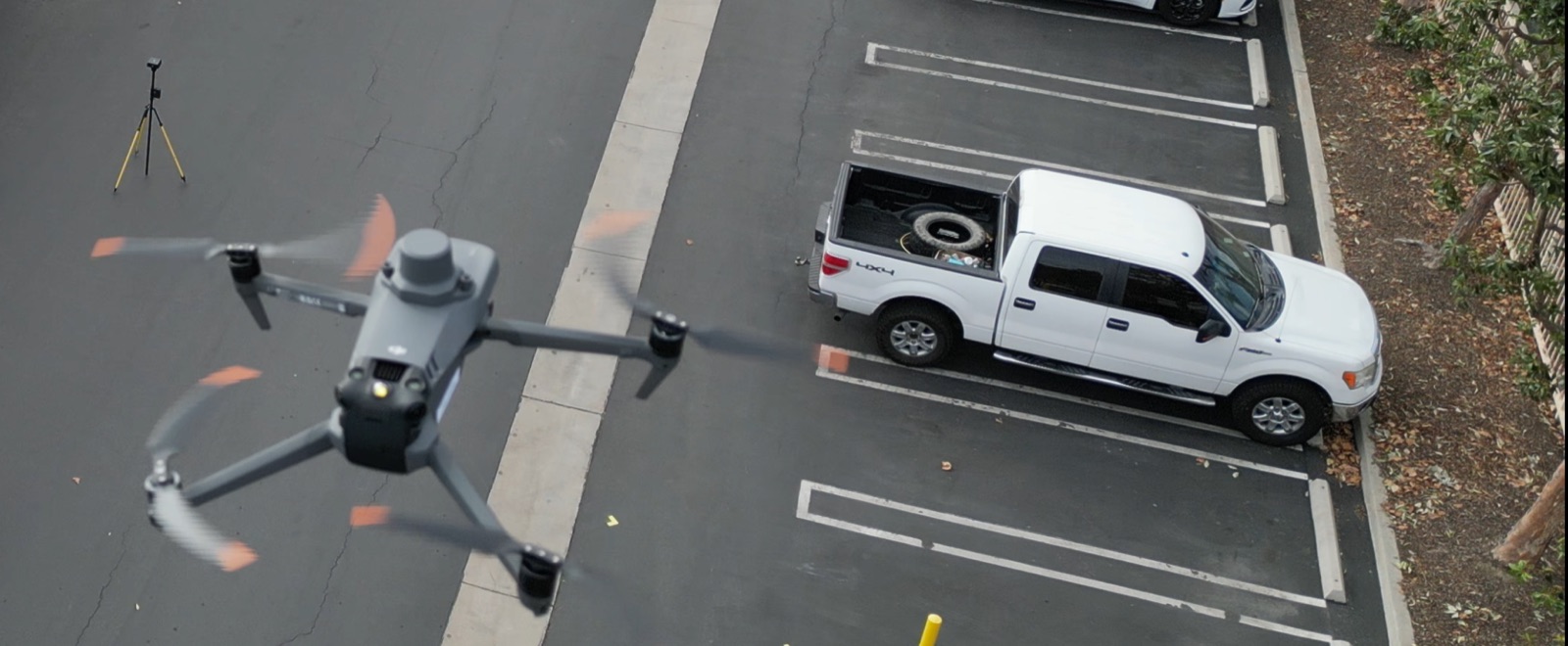 Drone flying above a paved site with a field checkpoint tripod and parked truck below.